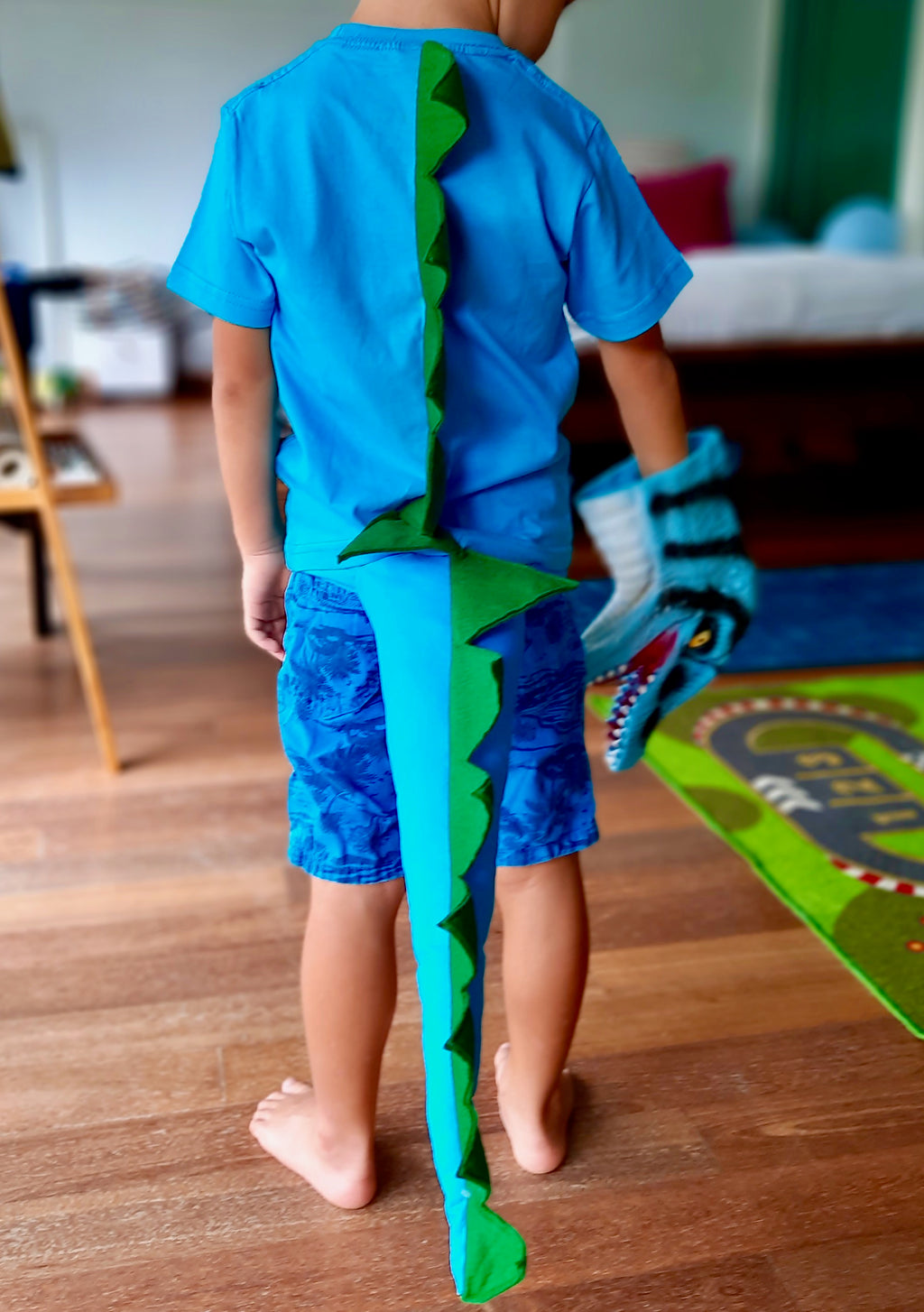 A child wearing a blue dinosaur costume with a green and blue tail, standing in a room with a colorful floor covering.