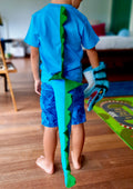 A child wearing a blue dinosaur costume with a green and blue tail, standing in a room with a colorful floor covering.