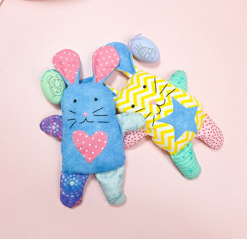 Two plush toy rabbits with colorful patterns, one in blue with a pink heart and polka dots, and the other in yellow with a geometric pattern.