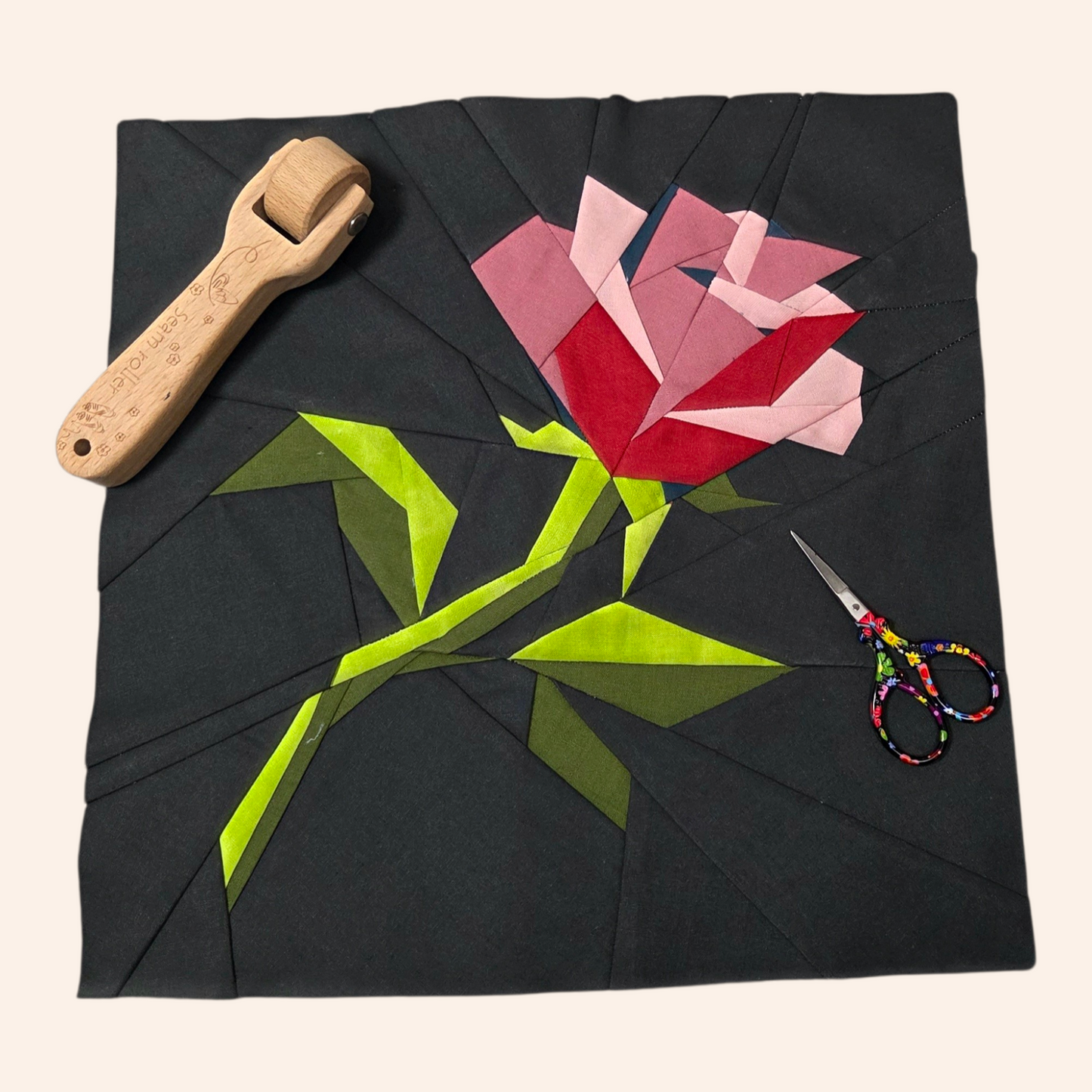 Quilt block with a red rose design on black fabric, accompanied by a wooden roller and scissors.