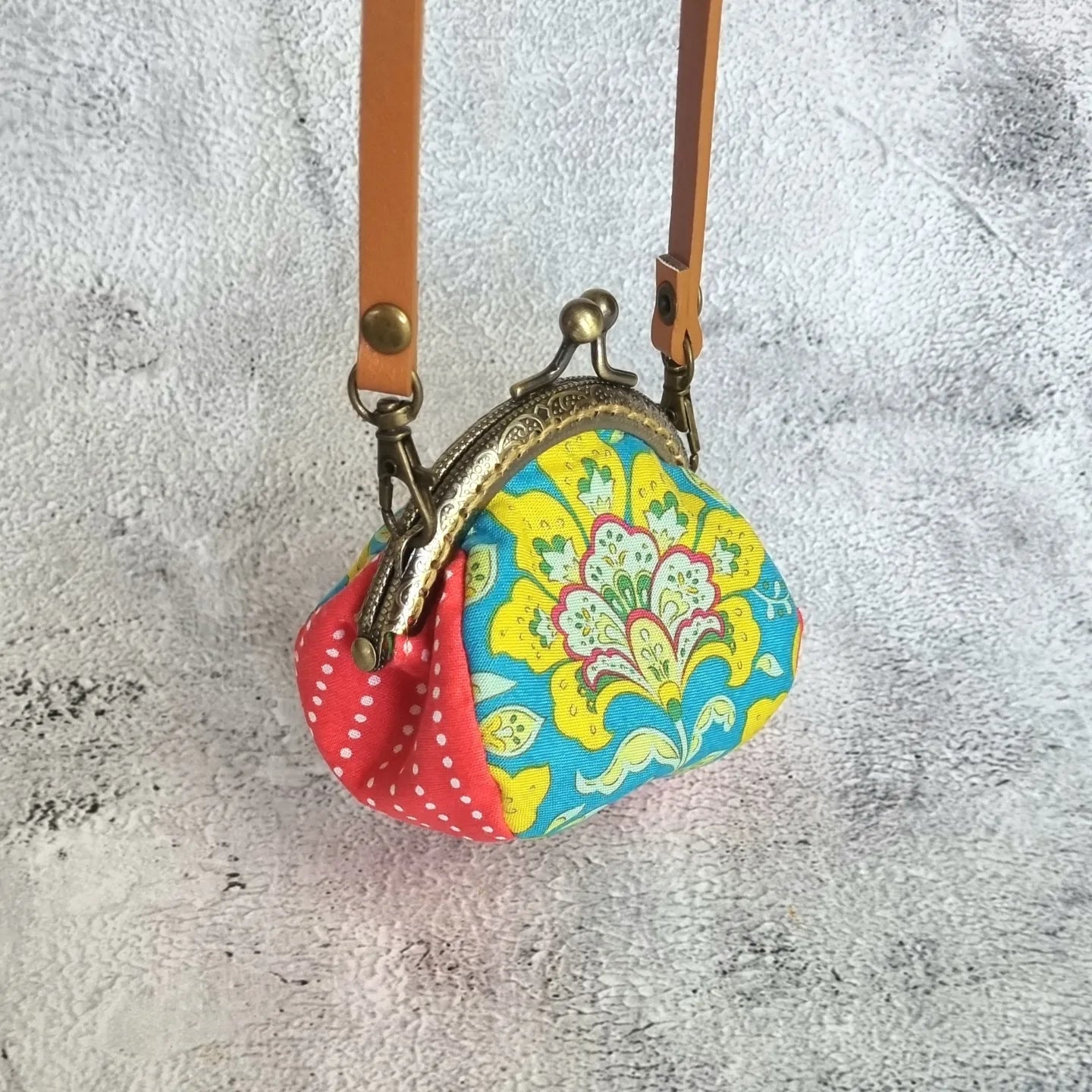 A handcrafted clasp purse with a red polka-dot pattern on the front and a floral pattern on the clasp, hanging from two leather straps.