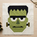 A Frankenstein-themed quilt block featuring olive green, black, and cream fabric panels, displayed on a light wood table with sewing scissors and thread spools.