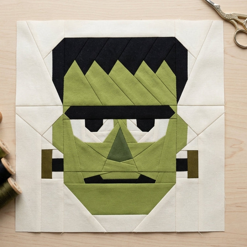 A Frankenstein-themed quilt block featuring olive green, black, and cream fabric panels, displayed on a light wood table with sewing scissors and thread spools.