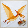 A quilted bird design made from geometric fabric pieces in orange, yellow, and rust tones on a white quilt, with three spools of thread and a pair of scissors placed in the lower right corner on a light wood surface.