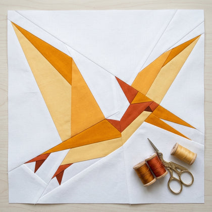 A quilted bird design made from geometric fabric pieces in orange, yellow, and rust tones on a white quilt, with three spools of thread and a pair of scissors placed in the lower right corner on a light wood surface.