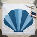 Quilt block with blue fan pattern on white fabric, scissors, and spools of thread on a wooden surface.