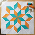 A quilted fabric block featuring a geometric starburst pattern in teal and orange on white, laid flat on a wooden surface with two spools of thread and a pair of scissors in the top right corner.
