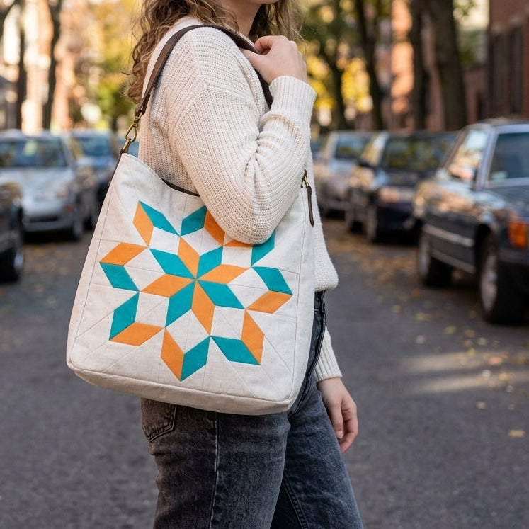 A woman stands on a city street carrying a large quilted tote bag. The bag is off-white with a colorful geometric starburst pattern in teal and orange.
