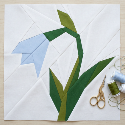 Quilt block with green and blue floral design on a white background, with sewing tools nearby.