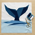 Quilt block with blue and white design on a wooden surface with scissors and thread spools.